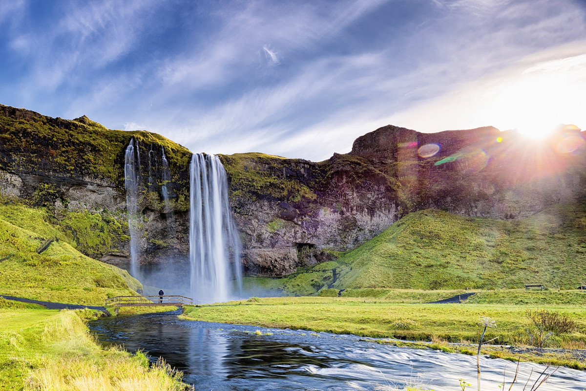 Ein majestätischer Wasserfall stürzt von einer moosbewachsenen Klippe in ein Flussbett. Im Vordergrund fließt ein Fluss durch eine grüne Ebene. Die Sonne scheint hell von rechts oben.